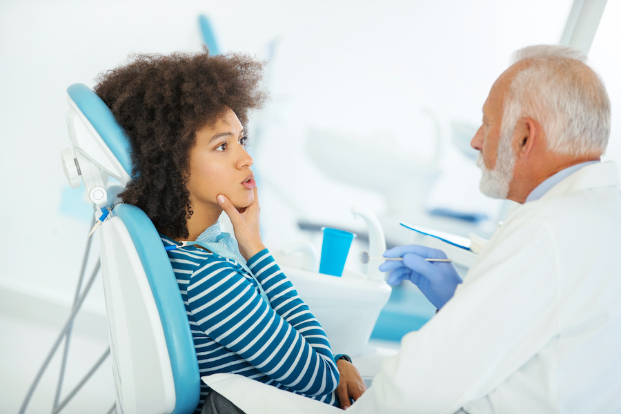 Curly-haired Black woman touches her jaw as her white older dentist explains a procedure to her