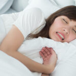 A brunette woman grinds and clenches her teeth as she sleeps in her white bedding