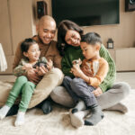 A mom and dad smile as they sit on the living room floor with their son and daughter playing dinosaurs
