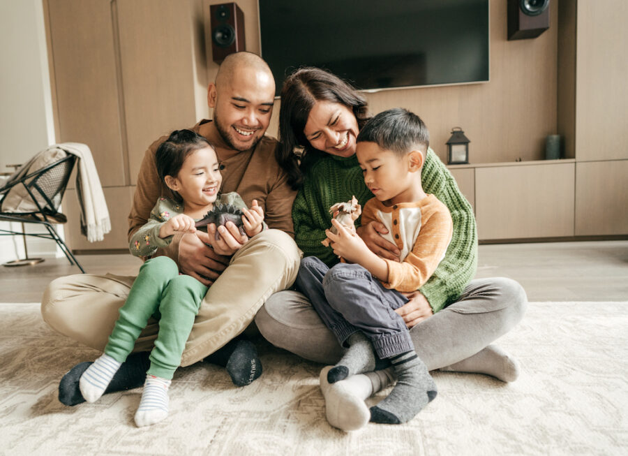 A mom and dad smile as they sit on the living room floor with their son and daughter playing dinosaurs