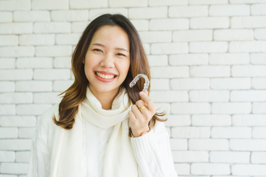 Asian woman in a cream turtleneck smiles while holding up her ClearCorrect clear aligners